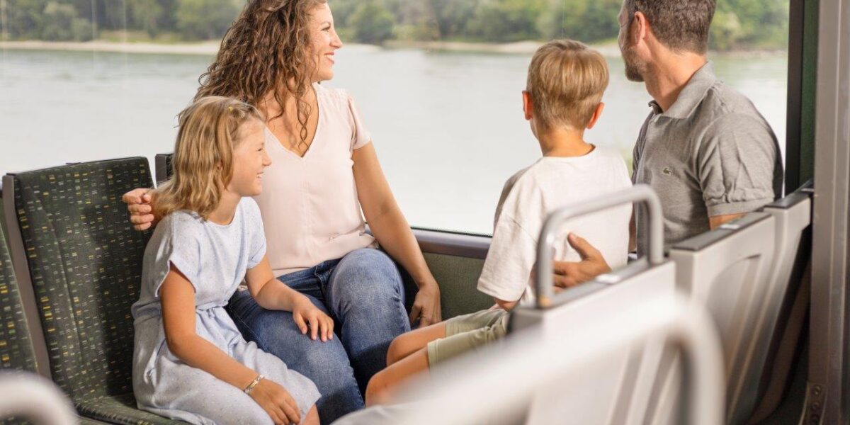 Eine vierköpfige Familie sitzt gemeinsam in einem Bus und blickt aus dem Fenster auf eine malerische Landschaft mit Wasser und Grünflächen.
