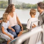 Eine vierköpfige Familie sitzt gemeinsam in einem Bus und blickt aus dem Fenster auf eine malerische Landschaft mit Wasser und Grünflächen.