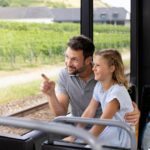 Ein Mann und ein junges Mädchen sitzen in einem Zug und schauen aus dem Fenster. Der Mann zeigt auf etwas, das draußen liegt, und die beiden scheinen beschäftigt zu sein. Ein Rucksack ruht auf dem Sitz neben ihnen.