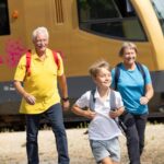 Ein Junge und zwei ältere Erwachsene mit Rucksäcken entfernen sich an einem sonnigen Tag von einem stehenden goldenen Zug.