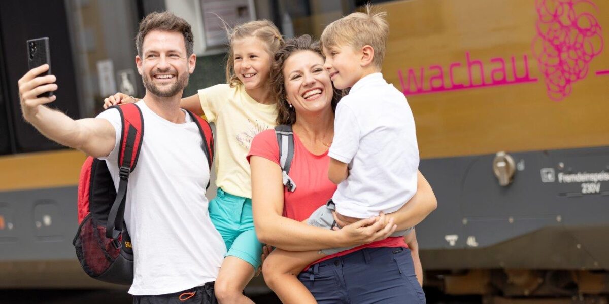 Eine vierköpfige Familie in Freizeitkleidung und mit Rucksäcken macht ein Selfie vor einem Zug, auf dessen Seite das Wort "Wachau" und ein Traubenlogo zu sehen sind.