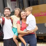 Eine vierköpfige Familie in Freizeitkleidung und mit Rucksäcken macht ein Selfie vor einem Zug, auf dessen Seite das Wort "Wachau" und ein Traubenlogo zu sehen sind.