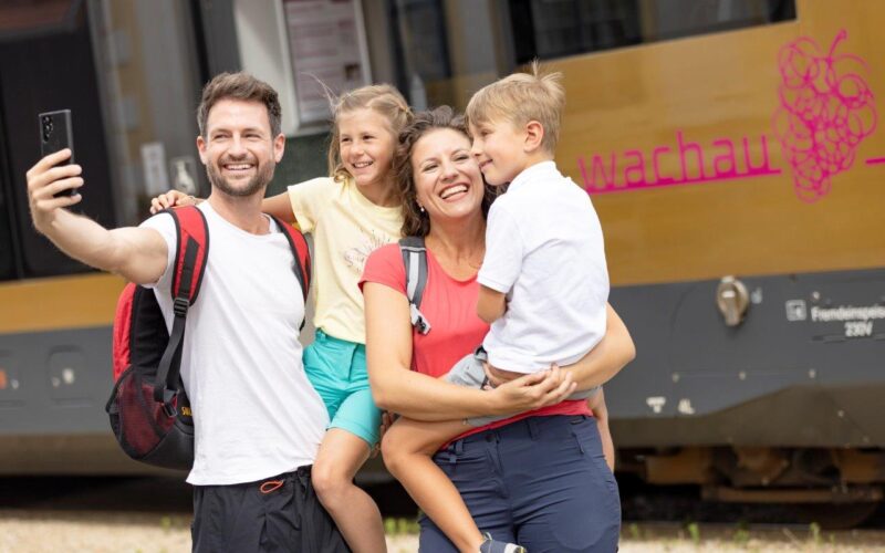 Eine vierköpfige Familie in Freizeitkleidung und mit Rucksäcken macht ein Selfie vor einem Zug, auf dessen Seite das Wort "Wachau" und ein Traubenlogo zu sehen sind.