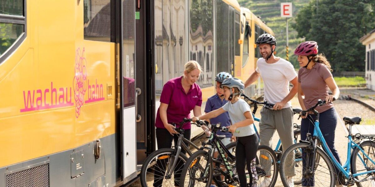 Zwei Erwachsene und zwei Kinder mit Fahrrädern bereiten sich darauf vor, in einen gelben Zug einzusteigen, während ein Mitarbeiter ihnen an einer Außenstation hilft.
