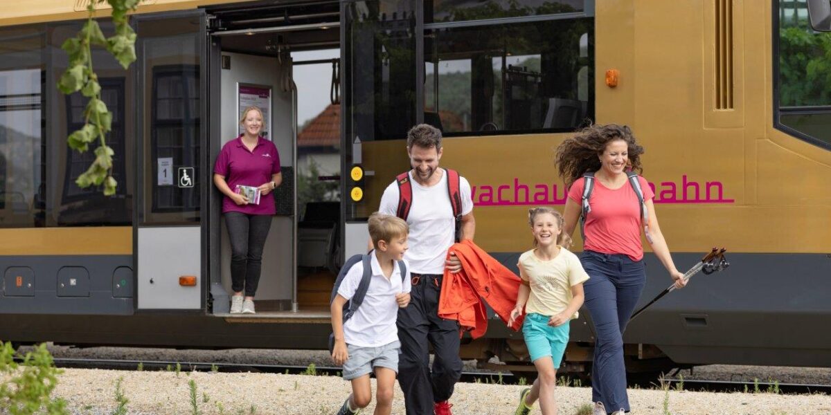 Eine Familie mit zwei Kindern verlässt einen stehenden Zug, während eine andere Frau lächelnd in der Tür des Zuges steht.