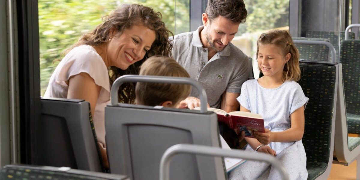 Eine Frau, ein Mann und zwei Kinder sitzen zusammen in einem Zug, wobei der Mann den Kindern ein Buch vorliest.