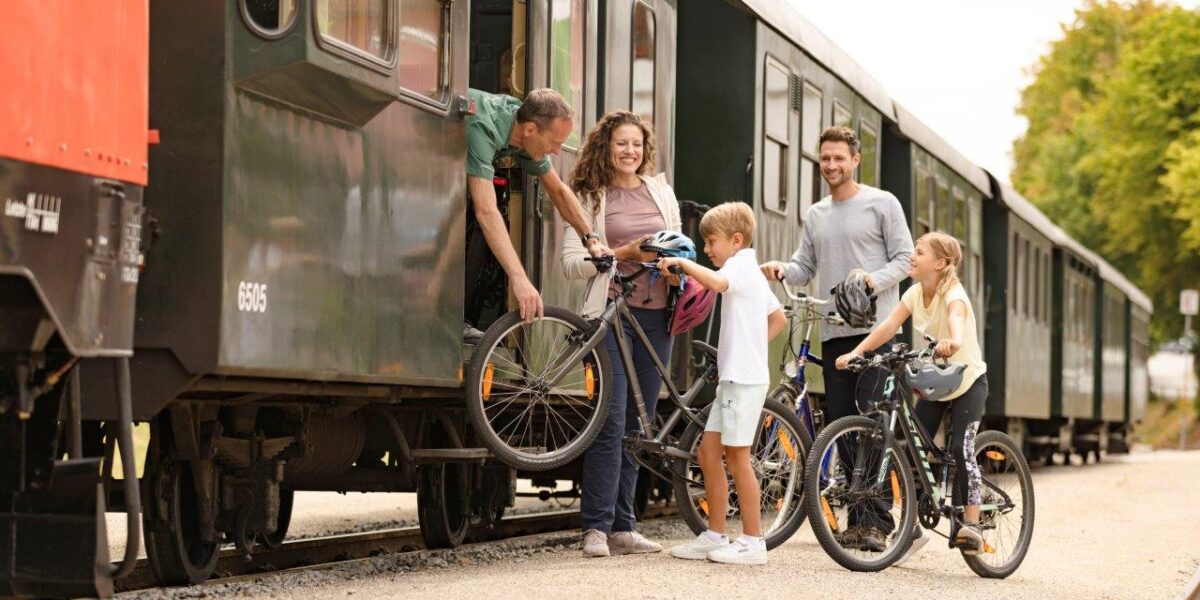 Eine Familie verlädt Fahrräder in einen stehenden Zug, während ein Bahnmitarbeiter ihr dabei hilft; zwei Kinder stehen mit ihren Fahrrädern auf dem Bahnsteig.