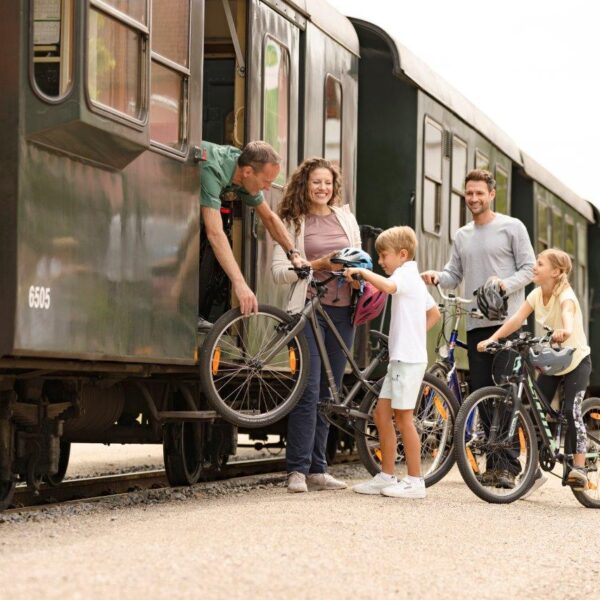 Eine Familie verlädt Fahrräder in einen stehenden Zug, während ein Bahnmitarbeiter ihr dabei hilft; zwei Kinder stehen mit ihren Fahrrädern auf dem Bahnsteig.