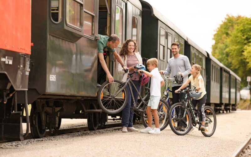 Eine Familie verlädt Fahrräder in einen stehenden Zug, während ein Bahnmitarbeiter ihr dabei hilft; zwei Kinder stehen mit ihren Fahrrädern auf dem Bahnsteig.