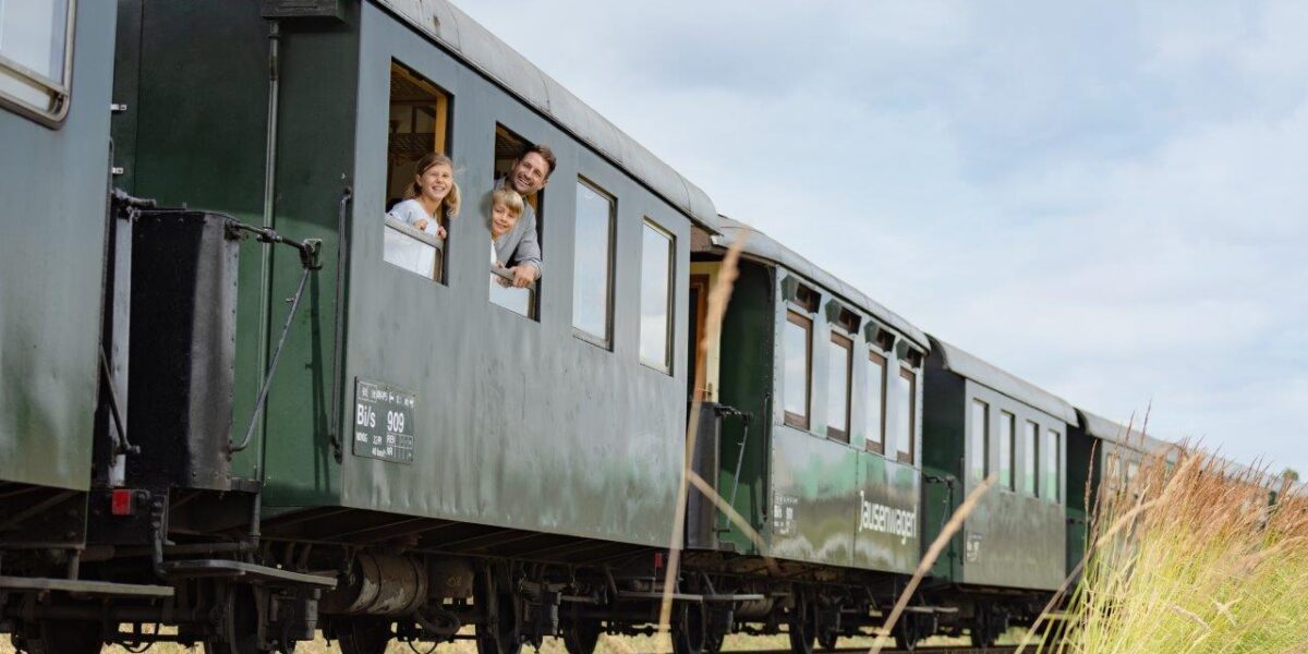 Drei Personen lehnen sich aus dem Fenster eines grünen Zugwagens, der auf Eisenbahnschienen durch eine grasbewachsene Landschaft fährt.