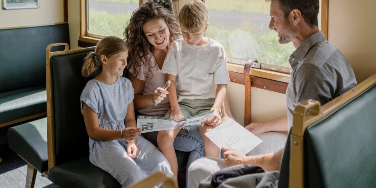 Eine vierköpfige Familie sitzt zusammen im Zug, die Kinder und die Mutter schauen sich eine Broschüre an, während der Vater lächelnd zusieht.