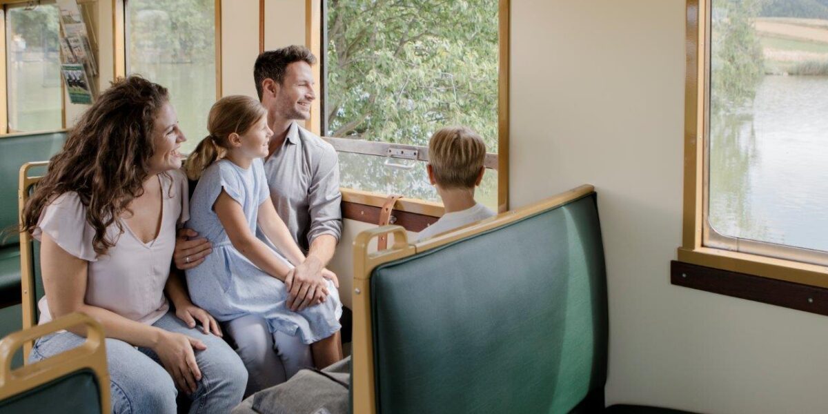 Eine vierköpfige Familie sitzt auf grünen Sitzen in einem Zug und blickt aus dem Fenster auf eine Seenlandschaft.