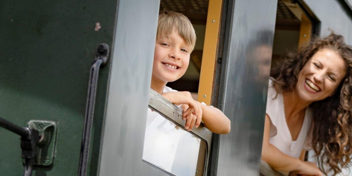 Ein kleiner Junge und eine Frau lehnen sich lächelnd aus dem Fenster eines grünen Zuges.