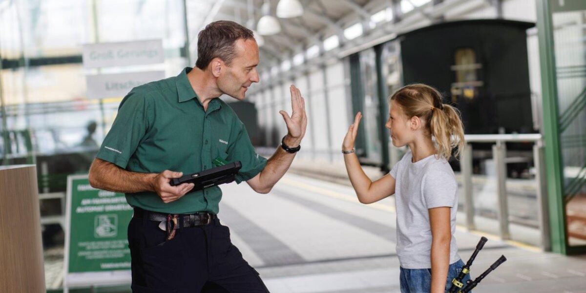 Ein Mann in einem grünen Hemd und ein junges Mädchen geben sich auf einem Bahnhof ein High Five, beide halten etwas in der Hand, das aussieht wie ein Modellbahnregler.