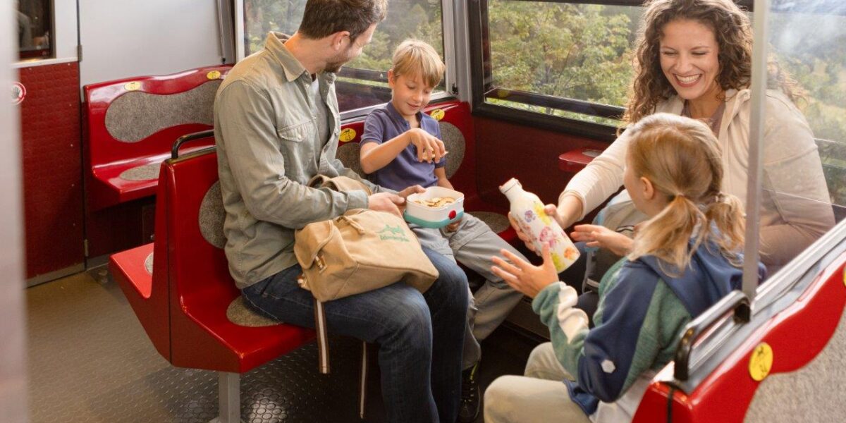 Ein Mann, eine Frau und zwei Kinder sitzen auf roten Sitzen in einem Zug oder einer Straßenbahn und teilen sich Snacks und Getränke an einem großen Fenster mit Blick auf die Landschaft.