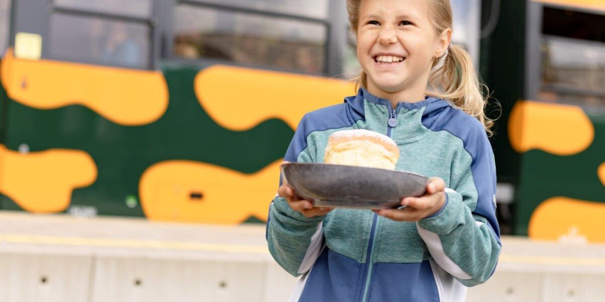 Ein lächelndes Kind in einer blauen Jacke hält einen Teller mit einem Sandwich und steht vor einem grün-gelben Zug.