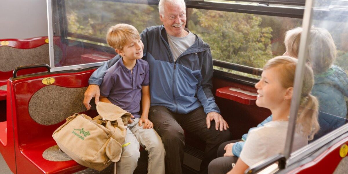 Ein älterer Mann und drei Kinder sitzen zusammen auf roten Sitzen in einem Zug oder einer Straßenbahn, unterhalten sich und lächeln mit Blick auf die Bäume vor dem Fenster.