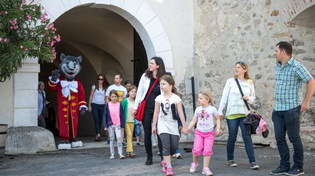 Eine Gruppe von Erwachsenen und Kindern geht an einer Person in einem Katzenmaskottchenkostüm vorbei, die in der Nähe eines gewölbten Steineingangs winkt.