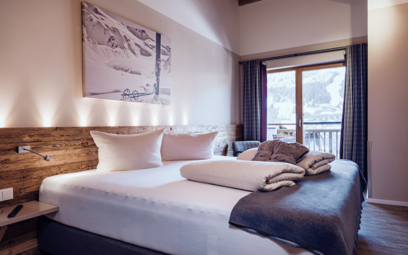 Modernes Hotelzimmer mit einem großen Bett, ordentlich gefalteten Handtüchern, einer Akzentwand aus Holz und einem Fenster mit Blick auf die verschneiten Berge draußen.