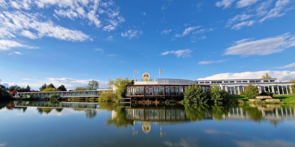 Ein Gebäude mit einer goldenen Löwenstatue auf dem Dach spiegelt sich in einem ruhigen See, umgeben von Bäumen und unter einem blauen Himmel mit vereinzelten Wolken.