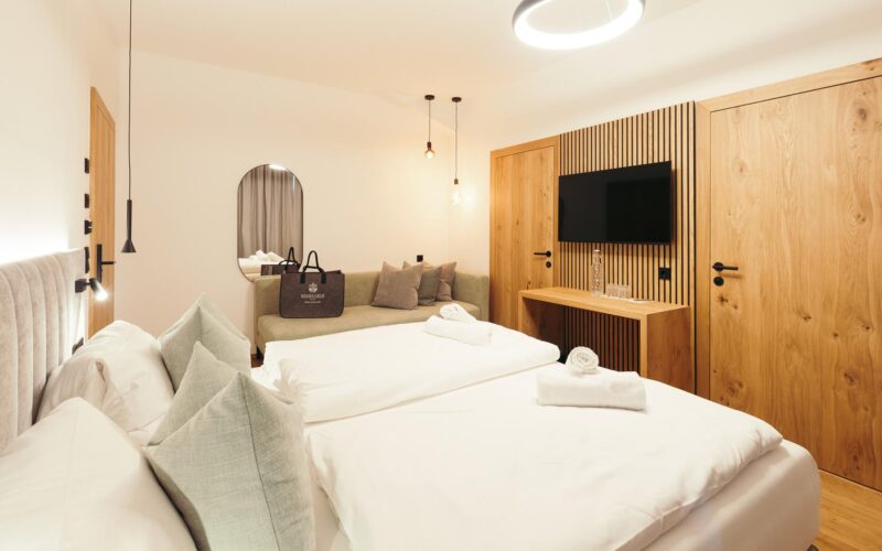 Modernes Hotelzimmer mit zwei sauber gemachten Einzelbetten, einem Sofa, einem an der Wand montierten Fernseher, Holzmöbeln und gedämpfter Umgebungsbeleuchtung.