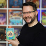 Ein Mann mit Brille und Bart lächelt in die Kamera und hält vor einem Regal voller Brettspiele ein buntes Veranstaltungsheft hoch.