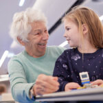 Eine ältere Frau und ein junges Mädchen sitzen zusammen im Haus, lächeln sich an, während sie an einem Tisch an einem kleinen Bauspielzeug arbeiten und ihr eigenes kleines Spielefest genießen.