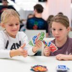 Zwei junge Mädchen sitzen an einem Tisch und spielen eine Partie UNO mit bunten Karten, im Hintergrund sind andere Leute zu sehen.