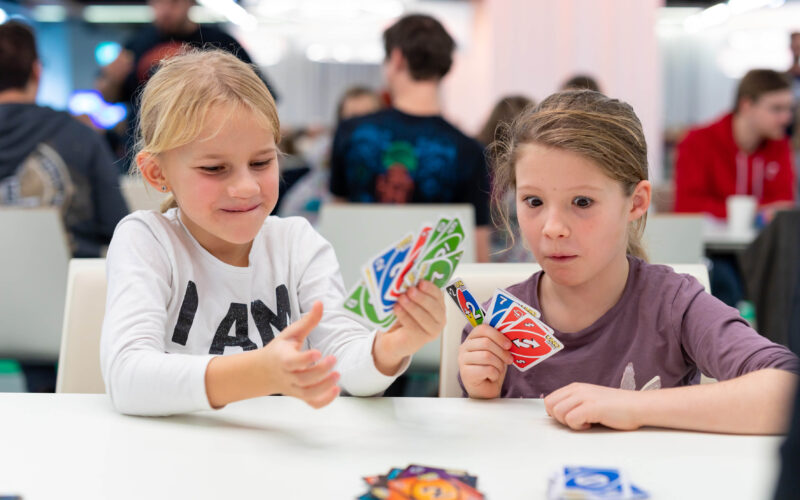 Zwei junge Mädchen sitzen an einem Tisch und spielen eine Partie UNO mit bunten Karten, im Hintergrund sind andere Leute zu sehen.