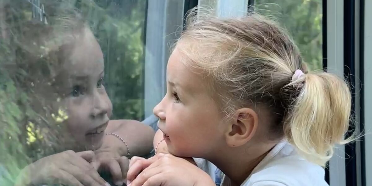 Ein junges Mädchen mit blondem Haar in einem Pferdeschwanz betrachtet ihr Spiegelbild in einem Fenster, aus dem Grünzeug zu sehen ist.