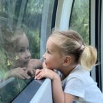 Ein junges Mädchen mit blondem Haar in einem Pferdeschwanz betrachtet ihr Spiegelbild in einem Fenster, aus dem Grünzeug zu sehen ist.