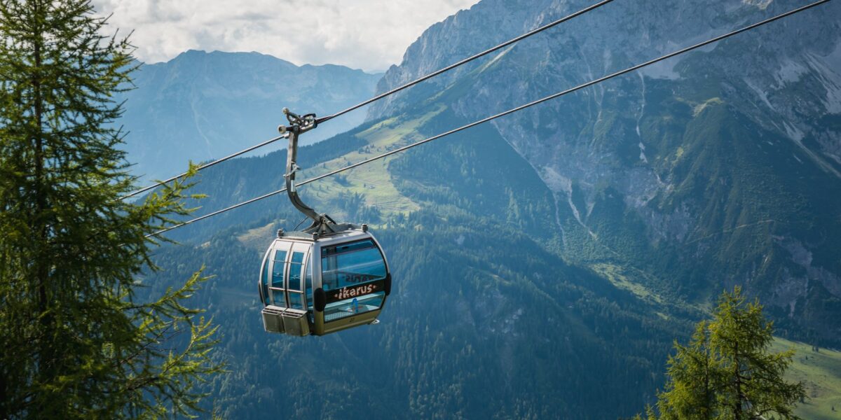 Eine blaue Gondelbahn fährt über grüne Bäume mit Bergen und Tälern im Hintergrund unter einem teilweise bewölkten Himmel.