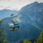 Eine blaue Gondelbahn fährt über grüne Bäume mit Bergen und Tälern im Hintergrund unter einem teilweise bewölkten Himmel.