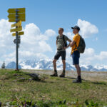 Zwei Personen mit Rucksäcken stehen in der Nähe eines gelben Wegweisers auf einer grasbewachsenen Hügelkuppe mit schneebedeckten Bergen und Wolken im Hintergrund.