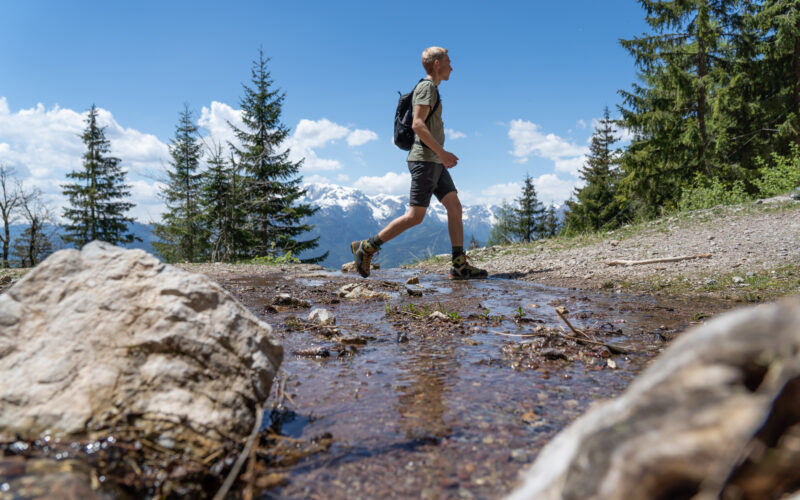 Eine Person mit einem Rucksack wandert auf einem felsigen Bergpfad in der Nähe eines seichten Baches, mit Bäumen und schneebedeckten Gipfeln im Hintergrund.
