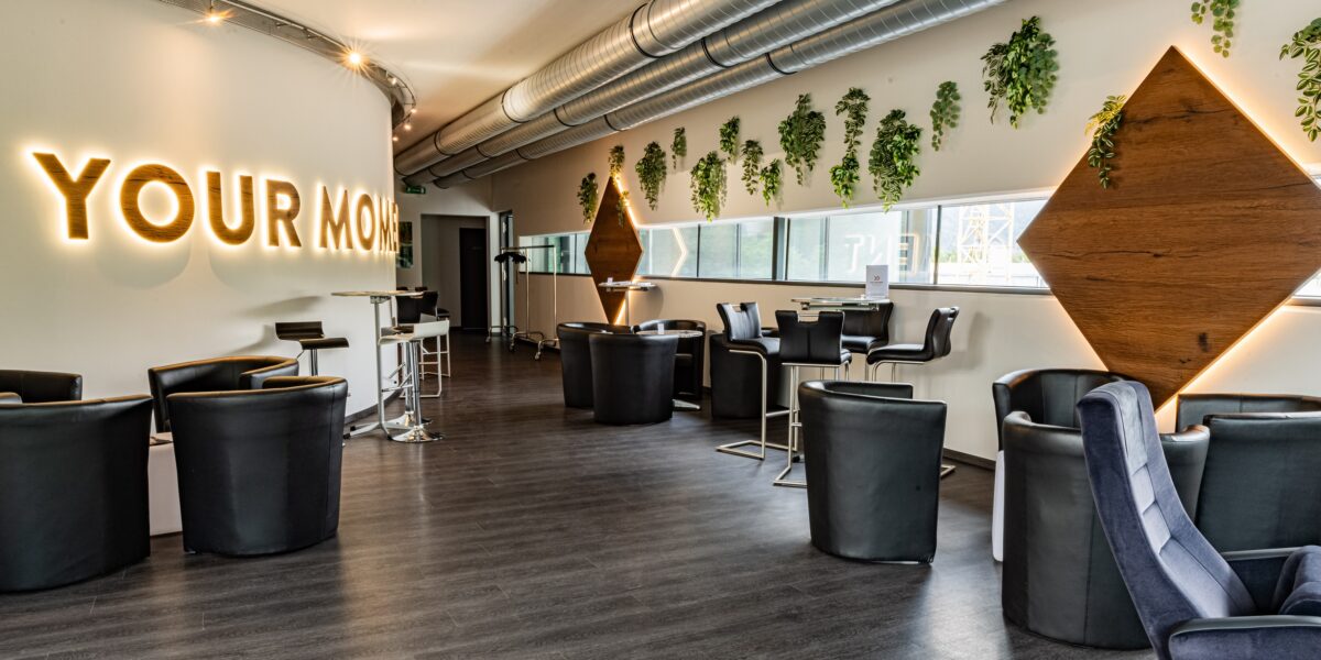 Moderner Lounge-Bereich mit schwarzen Stühlen, kleinen Tischen, Wandbegrünung, großen Rohrleitungen an der Decke und beleuchtetem "YOUR MOM"-Schild an einer gebogenen weißen Wand.