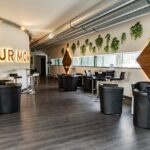 Moderner Lounge-Bereich mit schwarzen Stühlen, kleinen Tischen, Wandbegrünung, großen Rohrleitungen an der Decke und beleuchtetem "YOUR MOM"-Schild an einer gebogenen weißen Wand.