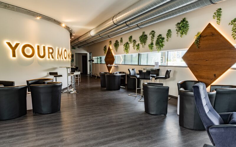 Moderner Lounge-Bereich mit schwarzen Stühlen, kleinen Tischen, Wandbegrünung, großen Rohrleitungen an der Decke und beleuchtetem "YOUR MOM"-Schild an einer gebogenen weißen Wand.