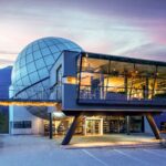 Ein modernes Gebäude mit einer großen kugelförmigen Struktur, einem verglasten Obergeschoss und Blick auf die Berge bei Sonnenuntergang; am Eingang sind Plakate und Lichter zu sehen.