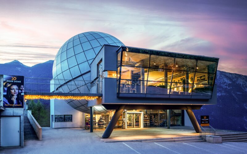 Ein modernes Gebäude mit einer großen kugelförmigen Struktur, einem verglasten Obergeschoss und Blick auf die Berge bei Sonnenuntergang; am Eingang sind Plakate und Lichter zu sehen.