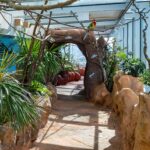 Ein Spazierweg mit künstlichen Steinen und Pflanzen, ein steinerner Torbogen und Modellpapageien und -echsen unter einem Glasdach in einem hell erleuchteten Innenraum.