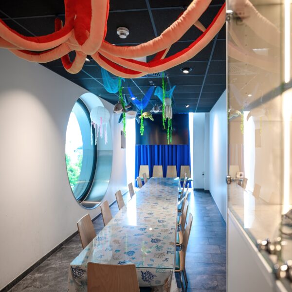 Ein Konferenzraum mit einem langen Tisch, Holzstühlen, großen runden Fenstern und hängenden Dekorationen mit Meeresmotiven an der Decke.