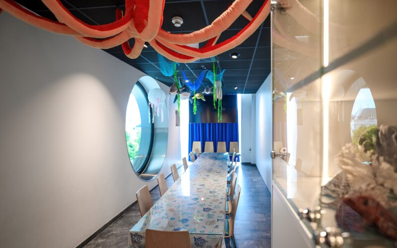 Ein Konferenzraum mit einem langen Tisch, Holzstühlen, großen runden Fenstern und hängenden Dekorationen mit Meeresmotiven an der Decke.