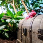 Zwischen grünen Pflanzen steht eine hölzerne Schatztruhe mit Metallschloss, auf der sich ein kleines rosa-rotes Plüschtier befindet.