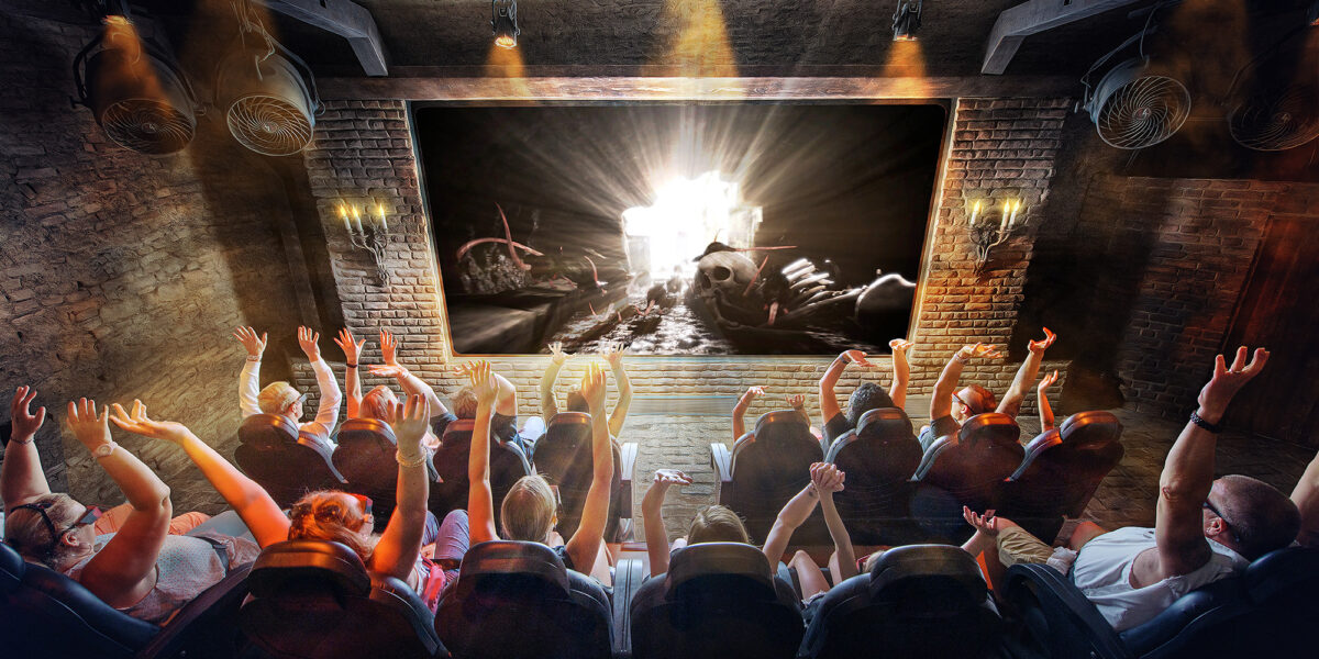 Eine Gruppe von Menschen sitzt mit erhobenen Armen in einem Theater und sieht sich eine helle, dramatische Szene auf einer großen Leinwand in einem schwach beleuchteten, industriell anmutenden Raum an.
