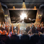 Eine Gruppe von Menschen sitzt mit erhobenen Armen in einem Theater und sieht sich eine helle, dramatische Szene auf einer großen Leinwand in einem schwach beleuchteten, industriell anmutenden Raum an.