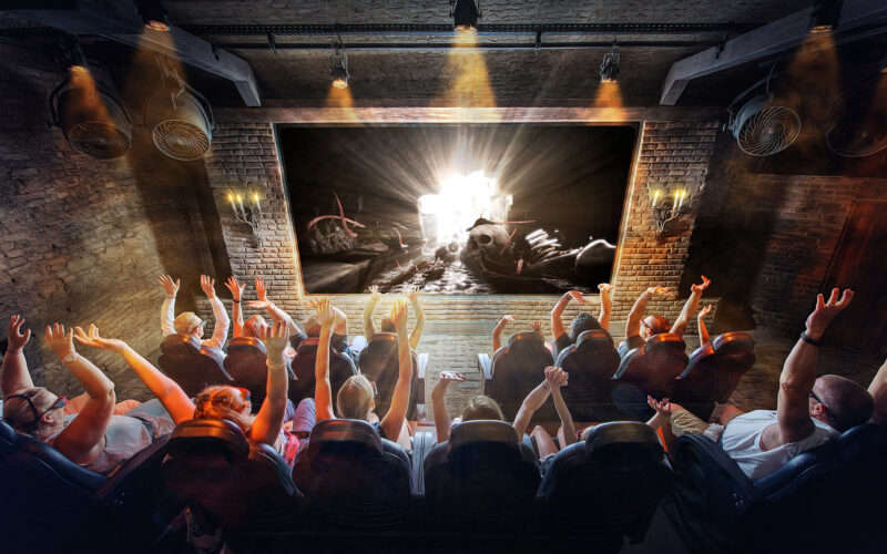 Eine Gruppe von Menschen sitzt mit erhobenen Armen in einem Theater und sieht sich eine helle, dramatische Szene auf einer großen Leinwand in einem schwach beleuchteten, industriell anmutenden Raum an.