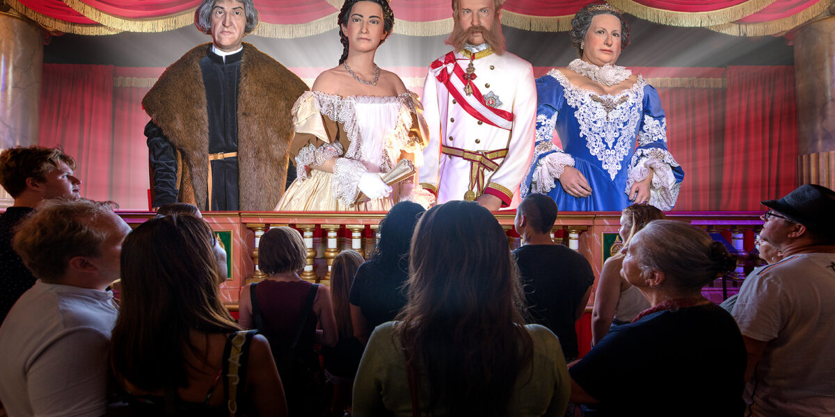 Eine Gruppe von Menschen steht vor einer Bühne, auf der vier lebensechte Wachsfiguren in aufwendigen historischen Kostümen bei theatralischer Beleuchtung stehen.