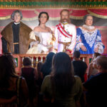 Eine Gruppe von Menschen steht vor einer Bühne, auf der vier lebensechte Wachsfiguren in aufwendigen historischen Kostümen bei theatralischer Beleuchtung stehen.