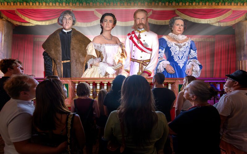 Eine Gruppe von Menschen steht vor einer Bühne, auf der vier lebensechte Wachsfiguren in aufwendigen historischen Kostümen bei theatralischer Beleuchtung stehen.